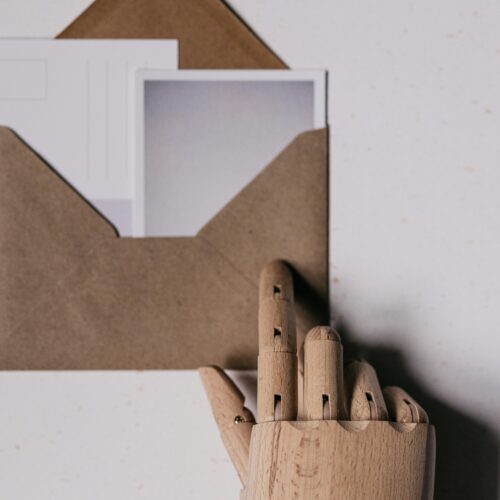 A wooden mannequin hand holds a brown envelope containing blank postcards.